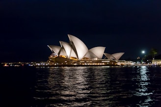 A night view of the Sydney Opera House, with its iconic white, sail-like structures illuminated against the dark sky. The reflection on the water adds to the picturesque scene, while city lights are visible in the distance.