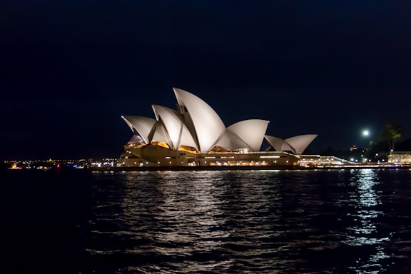 A night view of the Sydney Opera House, with its iconic white, sail-like structures illuminated against the dark sky. The reflection on the water adds to the picturesque scene, while city lights are visible in the distance.