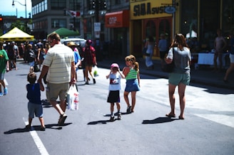People of various ages are walking along a street, presumably during a festival or a market event. There are shops and stalls with groups of people gathered around them. Some individuals are carrying shopping bags. The atmosphere suggests a busy and lively community event.