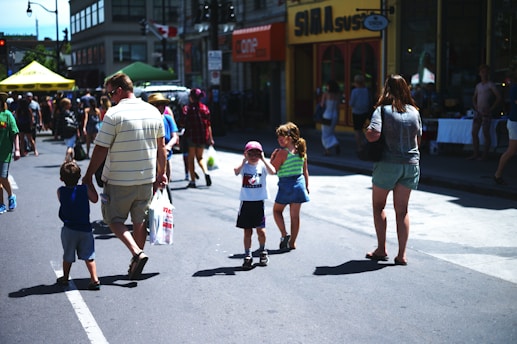 People of various ages are walking along a street, presumably during a festival or a market event. There are shops and stalls with groups of people gathered around them. Some individuals are carrying shopping bags. The atmosphere suggests a busy and lively community event.