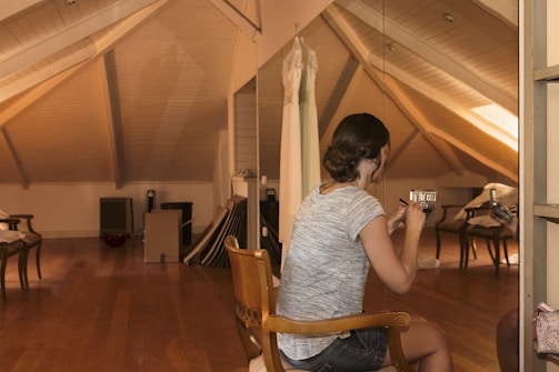 A woman is seated on a wooden chair in an attic room with sloped ceilings and wooden flooring. She appears to be applying makeup using a small mirror. The room is softly lit and includes a wedding dress hanging in the background. The environment is tidy, featuring mirrors, a few pieces of furniture, and some folded items stored against the walls.