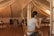 A woman is seated on a wooden chair in an attic room with sloped ceilings and wooden flooring. She appears to be applying makeup using a small mirror. The room is softly lit and includes a wedding dress hanging in the background. The environment is tidy, featuring mirrors, a few pieces of furniture, and some folded items stored against the walls.