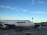A large commercial Qantas airplane is parked on an airport tarmac near a terminal building. The sky is clear with some scattered clouds, and the scene is lit with natural light from the setting or rising sun. Ground service vehicles are nearby, indicating preparation for flight operations.