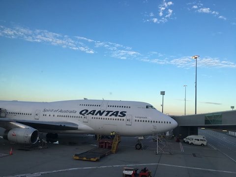 A large commercial Qantas airplane is parked on an airport tarmac near a terminal building. The sky is clear with some scattered clouds, and the scene is lit with natural light from the setting or rising sun. Ground service vehicles are nearby, indicating preparation for flight operations.