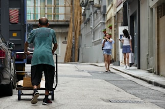 A man is pushing a cart filled with boxes down a narrow urban street. Two women are walking ahead, one looking at her phone and the other adjusting her hair. The street is lined with tall buildings and is relatively empty.