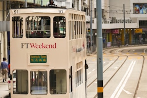 A vintage double-decker tram, painted in cream with 'FT Weekend' branding, travels on tracks through a city street. The tram displays 'Kennedy Town' as its destination. Surrounding the tram, various buildings line the street with signage written in Chinese characters. A few pedestrians walk nearby, and the tracks curve into the distance.