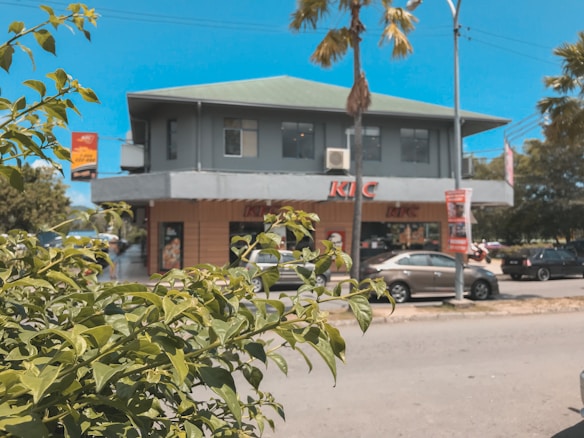 A commercial building with a fast-food restaurant on the ground floor is seen in a sunny outdoor setting. The building has a green roof and grey walls. In the foreground, leafy green shrubs are visible, and there are several parked cars near the building. The sky is clear and blue, with a few palm trees lining the road.