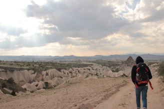 A person wearing a hoodie and a large backpack walks along a dirt path with a vast, rugged landscape in the background. The eroded rock formations known as fairy chimneys are visible, and the sky is partly cloudy with sun rays breaking through.