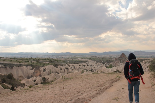 A person wearing a hoodie and a large backpack walks along a dirt path with a vast, rugged landscape in the background. The eroded rock formations known as fairy chimneys are visible, and the sky is partly cloudy with sun rays breaking through.