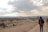 A person wearing a hoodie and a large backpack walks along a dirt path with a vast, rugged landscape in the background. The eroded rock formations known as fairy chimneys are visible, and the sky is partly cloudy with sun rays breaking through.