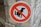 A circular sign with a white background and a red border, featuring a black silhouette of a dog squatting with a red diagonal line through it, indicating a prohibition. The sign is mounted on a metal pole against a stone wall background.