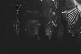 A black and white image of a live music performance, featuring a musician playing a guitar on stage. The stage is equipped with several lights and a large backdrop with a swirl design. A crowd can be seen in the foreground, with some hands raised, suggesting an engaging and lively concert atmosphere.