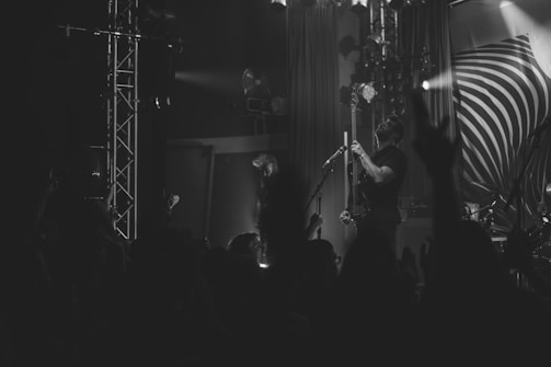 A black and white image of a live music performance, featuring a musician playing a guitar on stage. The stage is equipped with several lights and a large backdrop with a swirl design. A crowd can be seen in the foreground, with some hands raised, suggesting an engaging and lively concert atmosphere.