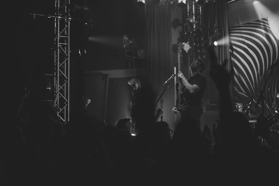 A black and white image of a live music performance, featuring a musician playing a guitar on stage. The stage is equipped with several lights and a large backdrop with a swirl design. A crowd can be seen in the foreground, with some hands raised, suggesting an engaging and lively concert atmosphere.