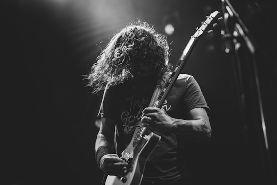 A black and white photo of a musician passionately playing an electric guitar. Their long hair is caught mid-swing, emphasizing the energy of the performance. The background is dimly lit, focusing attention on the musician's intense expression and the guitar.
