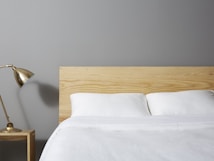 A minimalist bedroom scene features a neatly made bed with white linens and a light wooden headboard. A small wooden nightstand is placed beside the bed, supporting a stylish brass desk lamp. The background wall is painted in a soft gray color.