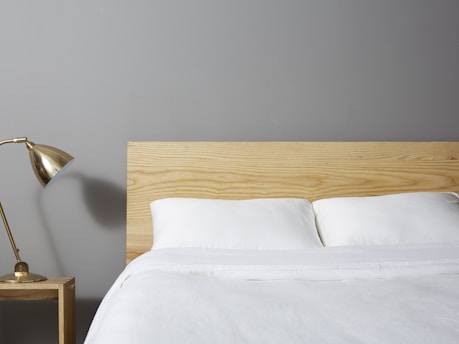 A minimalist bedroom scene features a neatly made bed with white linens and a light wooden headboard. A small wooden nightstand is placed beside the bed, supporting a stylish brass desk lamp. The background wall is painted in a soft gray color.