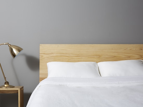 A minimalist bedroom scene features a neatly made bed with white linens and a light wooden headboard. A small wooden nightstand is placed beside the bed, supporting a stylish brass desk lamp. The background wall is painted in a soft gray color.