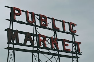 A large vintage-style sign with bold, red neon lettering spelling out 'PUBLIC MARKET' against a cloudy gray sky.