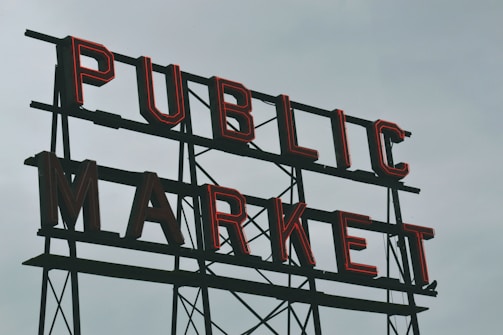 A large vintage-style sign with bold, red neon lettering spelling out 'PUBLIC MARKET' against a cloudy gray sky.
