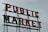 A large vintage-style sign with bold, red neon lettering spelling out 'PUBLIC MARKET' against a cloudy gray sky.