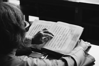 A person is reading a book filled with Chinese characters. The focus is on the hands holding the book and the detailed text on the pages. The setting appears calm and possibly within a study or library environment.