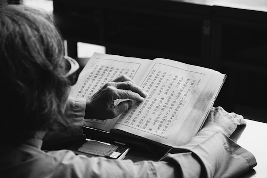 A person is reading a book filled with Chinese characters. The focus is on the hands holding the book and the detailed text on the pages. The setting appears calm and possibly within a study or library environment.
