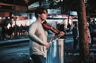 A person is playing a violin on a busy street at night. The scene is bustling with people and city lights, creating a lively urban atmosphere. The musician is focused, adding a sense of artistry to the urban setting.