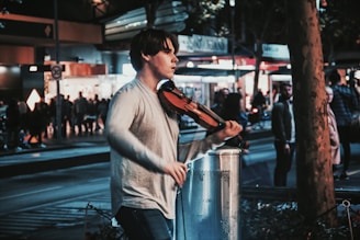 A person is playing a violin on a busy street at night. The scene is bustling with people and city lights, creating a lively urban atmosphere. The musician is focused, adding a sense of artistry to the urban setting.