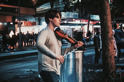 A person is playing a violin on a busy street at night. The scene is bustling with people and city lights, creating a lively urban atmosphere. The musician is focused, adding a sense of artistry to the urban setting.