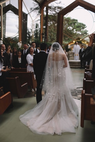 A groom wiping a tear as he watches his bride walk down the aisle in a sunlit chapel.