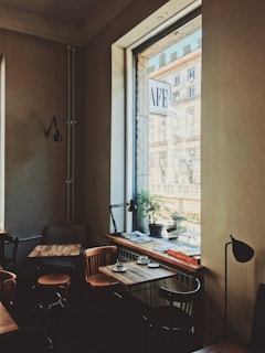 A cozy cafe interior with wooden tables and chairs arranged neatly. Large windows allow natural light to illuminate the space, revealing a street view outside. Some tables are adorned with cups and small plates, and there's a potted plant on the windowsill. The decor features minimalistic lamps and a warm, inviting color palette.