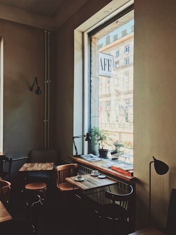 A cozy cafe interior with wooden tables and chairs arranged neatly. Large windows allow natural light to illuminate the space, revealing a street view outside. Some tables are adorned with cups and small plates, and there's a potted plant on the windowsill. The decor features minimalistic lamps and a warm, inviting color palette.