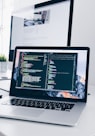 A MacBook with lines of code on its screen on a busy desk