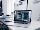 A MacBook with lines of code on its screen on a busy desk