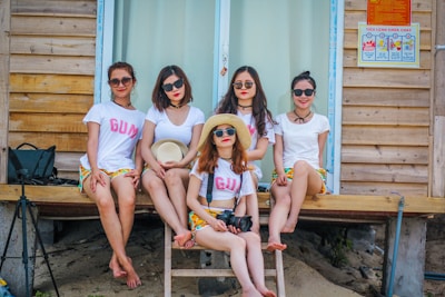 Five women are sitting on wooden steps in front of a cabin, all wearing sunglasses and white shirts with colorful shorts. One woman holds a hat, while another has a camera. The setting gives a casual, summery vibe.
