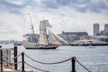 A large sailing ship with majestic sails is seen moving across a harbor, flying the American flag. The background features an urban skyline with tall buildings and a busy waterfront, exhibiting a mix of modern and older architecture. The scene is calm and somewhat overcast, with a small motorboat visible nearby.
