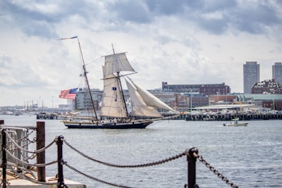 A large sailing ship with majestic sails is seen moving across a harbor, flying the American flag. The background features an urban skyline with tall buildings and a busy waterfront, exhibiting a mix of modern and older architecture. The scene is calm and somewhat overcast, with a small motorboat visible nearby.