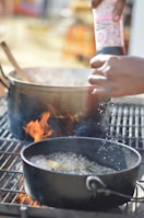 A hand is seasoning food in a cooking pot that is on a grill, with visible flames underneath. The scene suggests active cooking or grilling, with steam or bubbles visible on the surface of the contents.