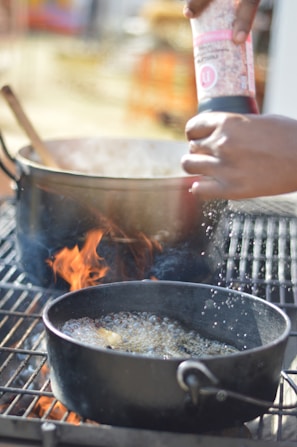 A hand is seasoning food in a cooking pot that is on a grill, with visible flames underneath. The scene suggests active cooking or grilling, with steam or bubbles visible on the surface of the contents.