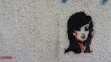 A stylized stencil painting of a woman's face with prominent black hair and red accents on a textured, light-colored wall. The artwork captures a mysterious and bold expression.
