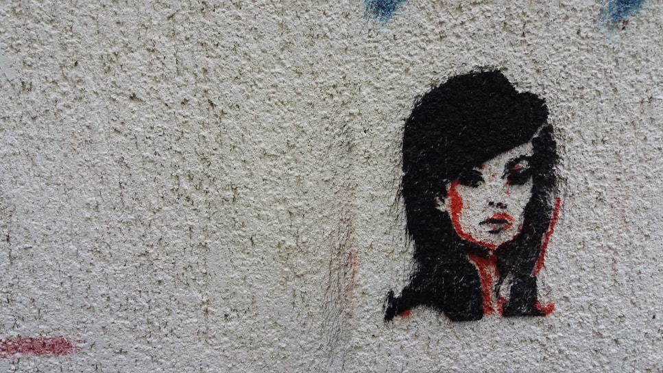 A stylized stencil painting of a woman's face with prominent black hair and red accents on a textured, light-colored wall. The artwork captures a mysterious and bold expression.