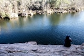 A person sits on a rocky cliff overlooking a calm body of water, with trees and shrubs lining the opposite shore. The water reflects some of the surroundings. The person appears to be alone and deep in thought.