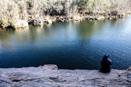 A person sits on a rocky cliff overlooking a calm body of water, with trees and shrubs lining the opposite shore. The water reflects some of the surroundings. The person appears to be alone and deep in thought.