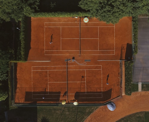An aerial view of a clay tennis court surrounded by lush green trees. Two players are seen on the courts, their shadows cast prominently on the vibrant orange surface. There are lines marking the boundaries of the court, and overhead netting partially visible.