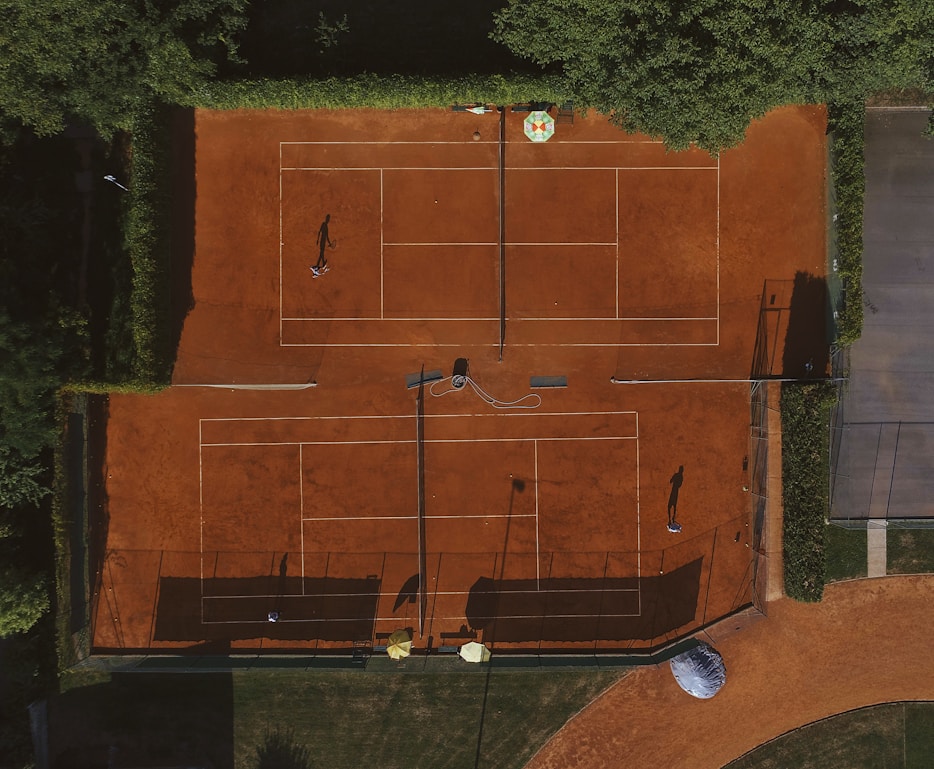An aerial view of a clay tennis court surrounded by lush green trees. Two players are seen on the courts, their shadows cast prominently on the vibrant orange surface. There are lines marking the boundaries of the court, and overhead netting partially visible.