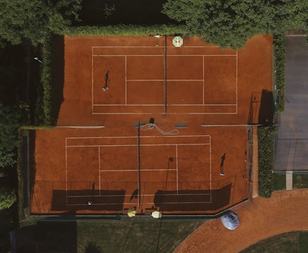 An aerial view of a clay tennis court surrounded by lush green trees. Two players are seen on the courts, their shadows cast prominently on the vibrant orange surface. There are lines marking the boundaries of the court, and overhead netting partially visible.