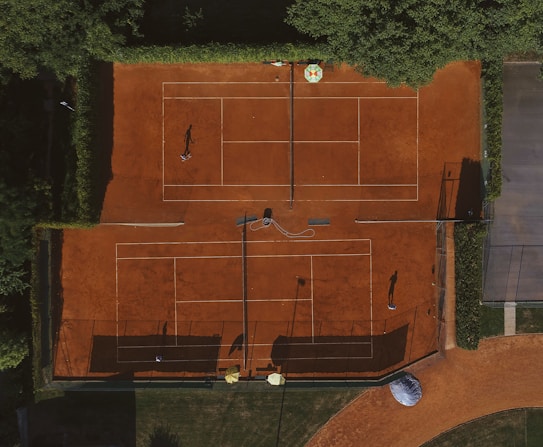 An aerial view of a clay tennis court surrounded by lush green trees. Two players are seen on the courts, their shadows cast prominently on the vibrant orange surface. There are lines marking the boundaries of the court, and overhead netting partially visible.