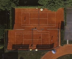 An aerial view of a clay tennis court surrounded by lush green trees. Two players are seen on the courts, their shadows cast prominently on the vibrant orange surface. There are lines marking the boundaries of the court, and overhead netting partially visible.