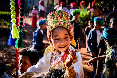 A young person wearing a colorful and ornate traditional costume with a bright headdress smiles amidst a lively outdoor gathering. Several people are dressed in vibrant clothing and head scarves in the background, suggesting a cultural or festive event. Sunlight creates shadows and highlights details in their clothing.